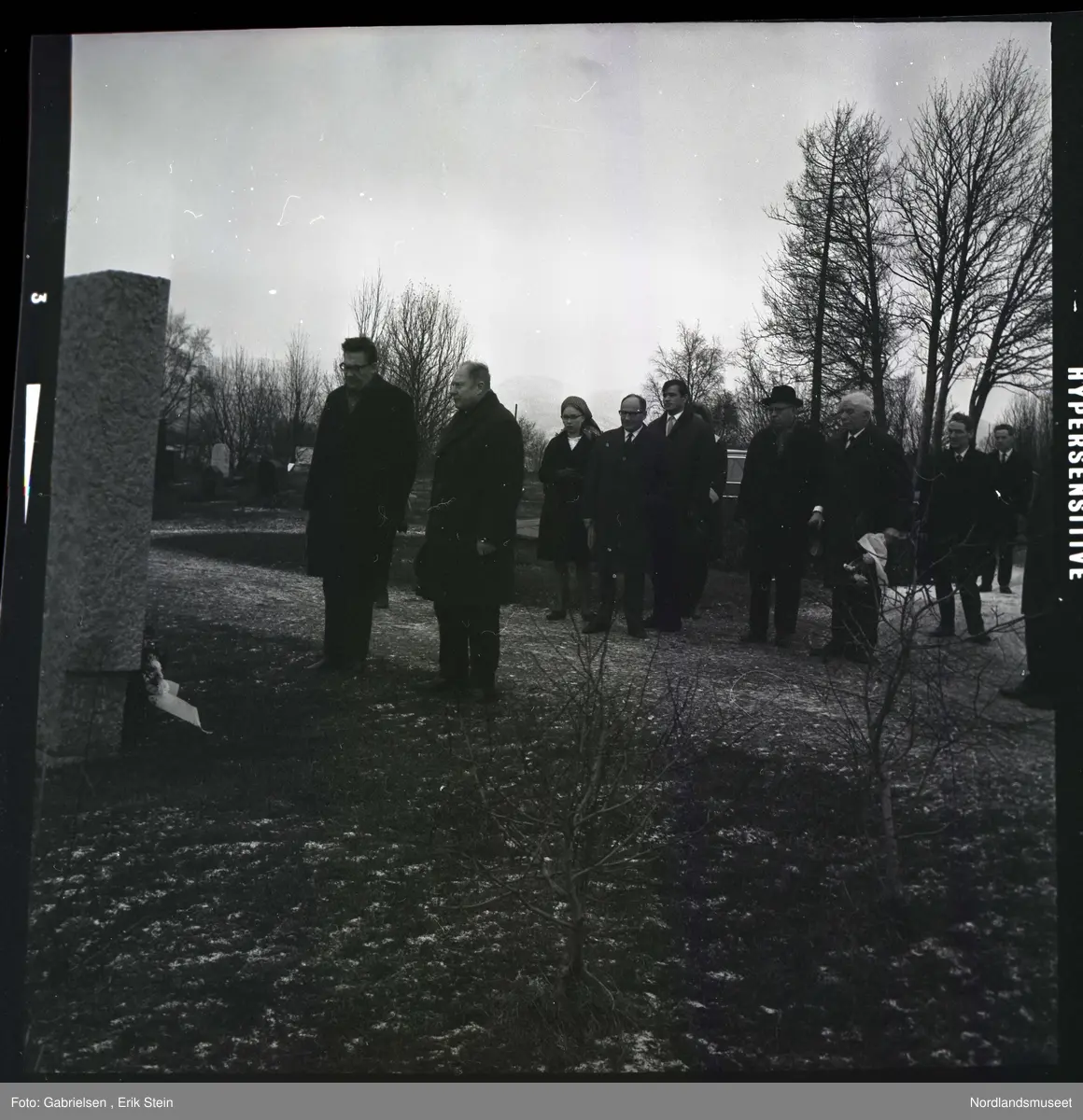 Fotografi av mange finkledde kvinner og menn i en minnemarkering på Fauske kirkegård som står og ser på en minnestein som de to fremste mennene har lagt en krans på 
til minne om falne soldater i Fauske under andre verdenskrig
og man ser en bil som står parkert på kirkegården
bak kvinnene og mennene vedsiden av noen trær 
som vokser på kirkegården og man ser to småtrær
som vokser i nærheten av minnesteinen 
og man ser en grusvei som går til kirkegården fra riksveien mot 
Bodø som går forbi kirkegården til Fauskekirken