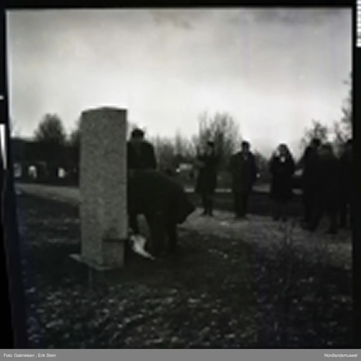 Fotografi av en minnemarkering på Fauske kirkegård 
som det står noen finkledde kvinner og menn og ser på to menn som legger ned en krans på en minnestein 
til falne soldater i Fauske under andre verdenskrig 
og en kvinne som tar bilde av mennen som legger ned kransen 
og man ser en fontene bak mennen og kvinnen i kirkegården
som det ligger et lokk over og man ser noen trær som vokser i kirkegården og en grusvei som går opp til kirkegården 
og mange gravsteiner i kirkegården