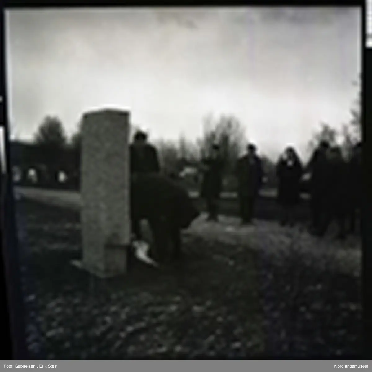 Fotografi av en minnemarkering på Fauske kirkegård 
som det står noen finkledde kvinner og menn og ser på to menn som legger ned en krans på en minnestein 
til falne soldater i Fauske under andre verdenskrig 
og en kvinne som tar bilde av mennen som legger ned kransen 
og man ser en fontene bak mennen og kvinnen i kirkegården
som det ligger et lokk over og man ser noen trær som vokser i kirkegården og en grusvei som går opp til kirkegården 
og mange gravsteiner i kirkegården