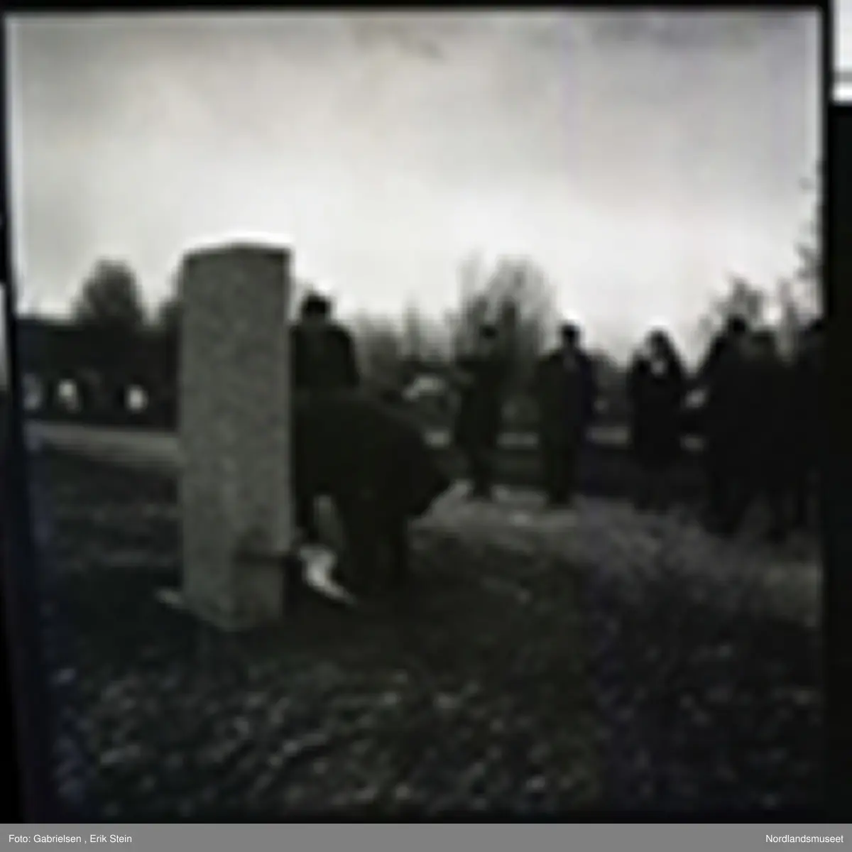 Fotografi av en minnemarkering på Fauske kirkegård 
som det står noen finkledde kvinner og menn og ser på to menn som legger ned en krans på en minnestein 
til falne soldater i Fauske under andre verdenskrig 
og en kvinne som tar bilde av mennen som legger ned kransen 
og man ser en fontene bak mennen og kvinnen i kirkegården
som det ligger et lokk over og man ser noen trær som vokser i kirkegården og en grusvei som går opp til kirkegården 
og mange gravsteiner i kirkegården