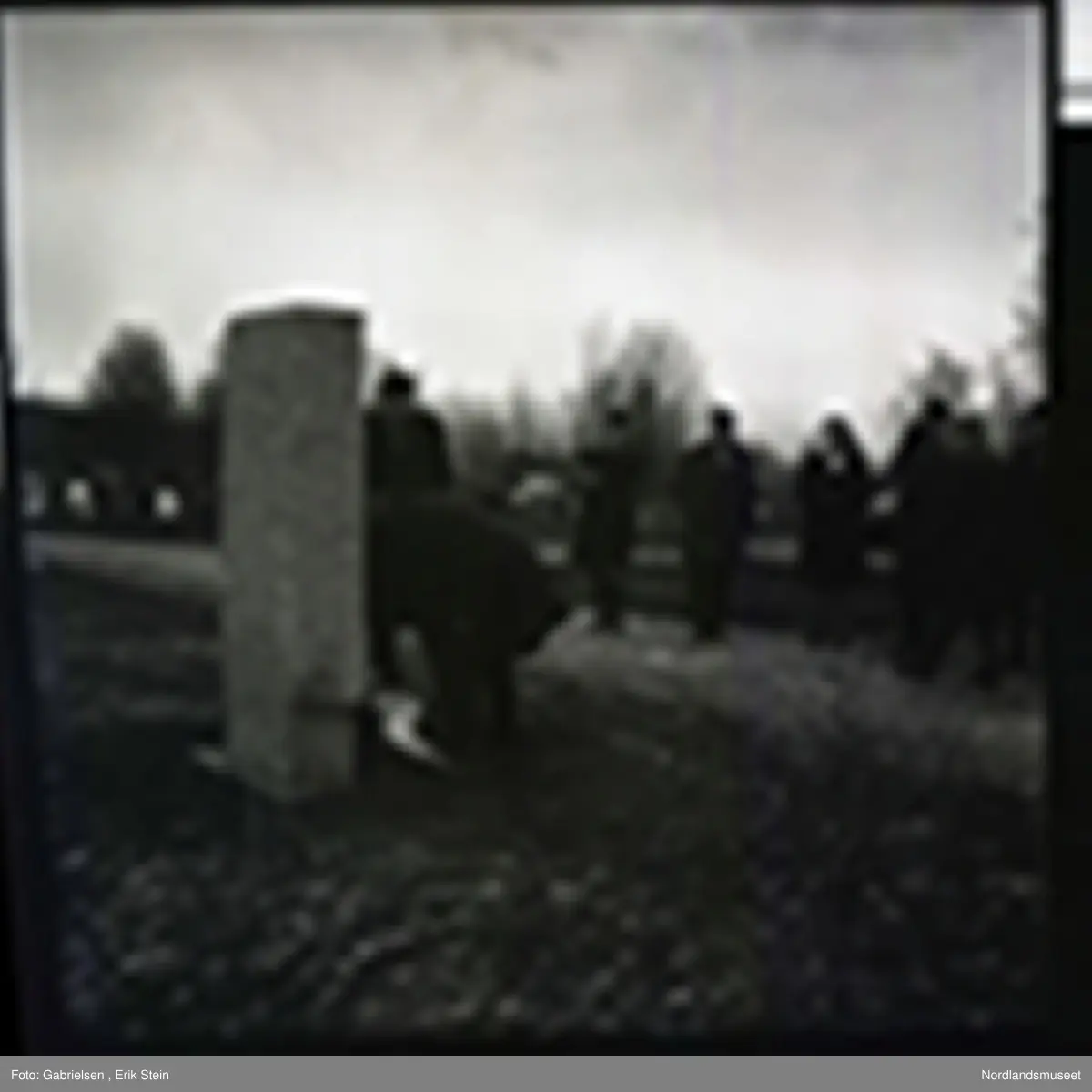 Fotografi av en minnemarkering på Fauske kirkegård 
som det står noen finkledde kvinner og menn og ser på to menn som legger ned en krans på en minnestein 
til falne soldater i Fauske under andre verdenskrig 
og en kvinne som tar bilde av mennen som legger ned kransen 
og man ser en fontene bak mennen og kvinnen i kirkegården
som det ligger et lokk over og man ser noen trær som vokser i kirkegården og en grusvei som går opp til kirkegården 
og mange gravsteiner i kirkegården