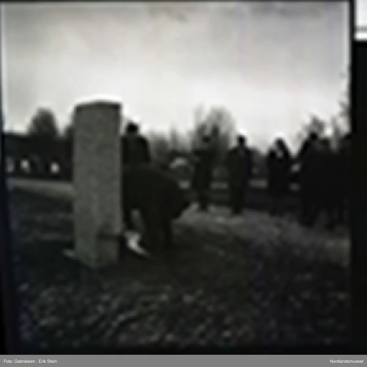 Fotografi av en minnemarkering på Fauske kirkegård 
som det står noen finkledde kvinner og menn og ser på to menn som legger ned en krans på en minnestein 
til falne soldater i Fauske under andre verdenskrig 
og en kvinne som tar bilde av mennen som legger ned kransen 
og man ser en fontene bak mennen og kvinnen i kirkegården
som det ligger et lokk over og man ser noen trær som vokser i kirkegården og en grusvei som går opp til kirkegården 
og mange gravsteiner i kirkegården
