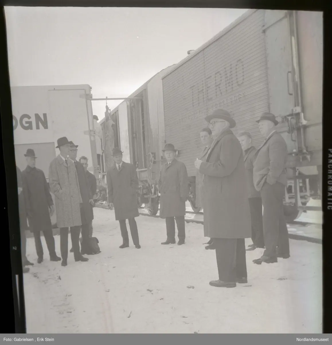 Fotografi av 11 menn kledd i dresser med slips
og hatter og jakker og andre klær 
som står vedsiden av et togvogn til et godstog
som man ser blir lastet på av en mann 
fra en lastebilvogn vedsiden av toget 
i en togstasjon et ukjent sted som det har snødd på
man ser at det ligger en koffert under en av mennene
i snøen og man ser to av mennene har briller på seg