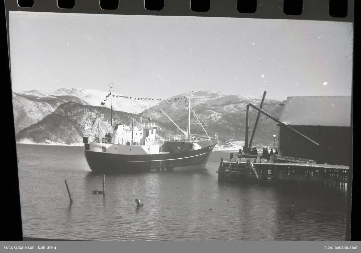 Fotografi av en båt som legger til fra Rognankaien
på sin jomfrutur igjennom Rognanfjorden mens noen personer som står på bryggen i kaien og ser på båten og man ser at båtens 
navn står på baugen og styrhuset til båten og 
to master som det er hengt et langt tau med mange 
forskjellige flagg og man ser en kran framme oppå
baugen til skipet og man ser noen rellinger som går langs akter og forsiden av båten og man ser noen personer som står på båten og ser mot kaien og man ser et båthus på kaien vedsiden av bryggen og noen tønner og noe ved som ligger på bryggen i kaien