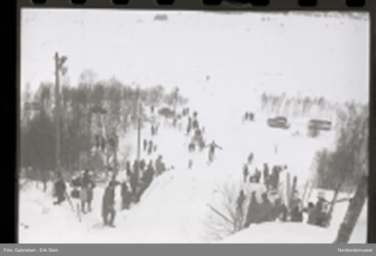 Fotografi av et skihopp som det hopper et barn med ski på seg fra mens noen kvinner og menn og barn med ski og skistaver og vinterklær og deltakernummer på seg som står og ser på barnet som hopper fra skihoppet vedsiden av skihoppet og lenger nede i bakken og tre barn som står i et utsiktstårn 
og ser på skihopperen og noen biler og busser som står parkert lenger nede i skibakken vedsiden av en vei som det vokser noen trær vedsiden av og man ser en skiløper ute i snømarken som har nettopp landet i snøen etter å ha hoppet fra skihoppet