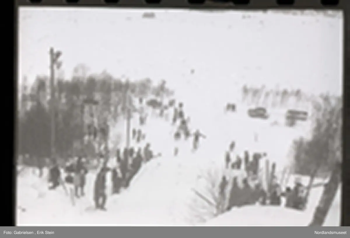 Fotografi av et skihopp som det hopper et barn med ski på seg fra mens noen kvinner og menn og barn med ski og skistaver og vinterklær og deltakernummer på seg som står og ser på barnet som hopper fra skihoppet vedsiden av skihoppet og lenger nede i bakken og tre barn som står i et utsiktstårn 
og ser på skihopperen og noen biler og busser som står parkert lenger nede i skibakken vedsiden av en vei som det vokser noen trær vedsiden av og man ser en skiløper ute i snømarken som har nettopp landet i snøen etter å ha hoppet fra skihoppet