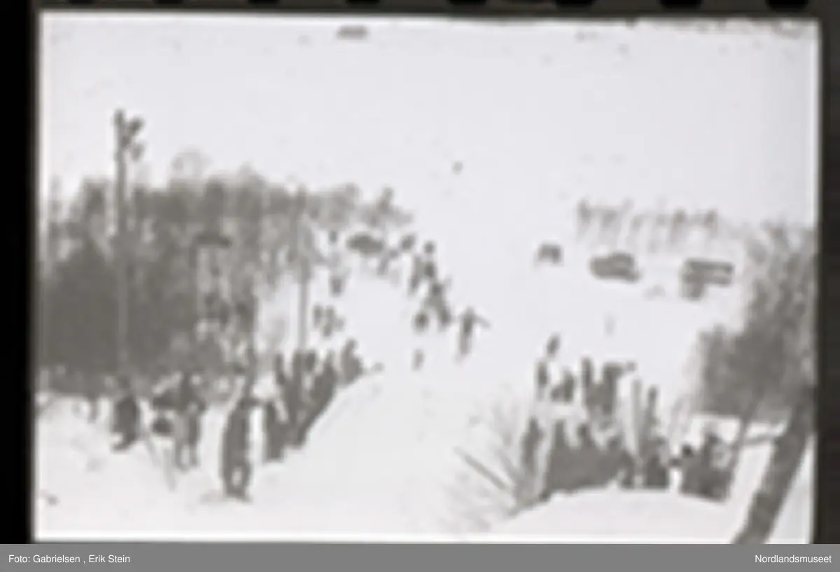 Fotografi av et skihopp som det hopper et barn med ski på seg fra mens noen kvinner og menn og barn med ski og skistaver og vinterklær og deltakernummer på seg som står og ser på barnet som hopper fra skihoppet vedsiden av skihoppet og lenger nede i bakken og tre barn som står i et utsiktstårn 
og ser på skihopperen og noen biler og busser som står parkert lenger nede i skibakken vedsiden av en vei som det vokser noen trær vedsiden av og man ser en skiløper ute i snømarken som har nettopp landet i snøen etter å ha hoppet fra skihoppet