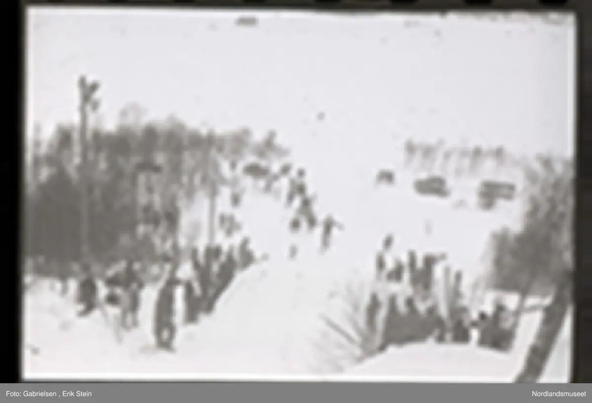 Fotografi av et skihopp som det hopper et barn med ski på seg fra mens noen kvinner og menn og barn med ski og skistaver og vinterklær og deltakernummer på seg som står og ser på barnet som hopper fra skihoppet vedsiden av skihoppet og lenger nede i bakken og tre barn som står i et utsiktstårn 
og ser på skihopperen og noen biler og busser som står parkert lenger nede i skibakken vedsiden av en vei som det vokser noen trær vedsiden av og man ser en skiløper ute i snømarken som har nettopp landet i snøen etter å ha hoppet fra skihoppet
