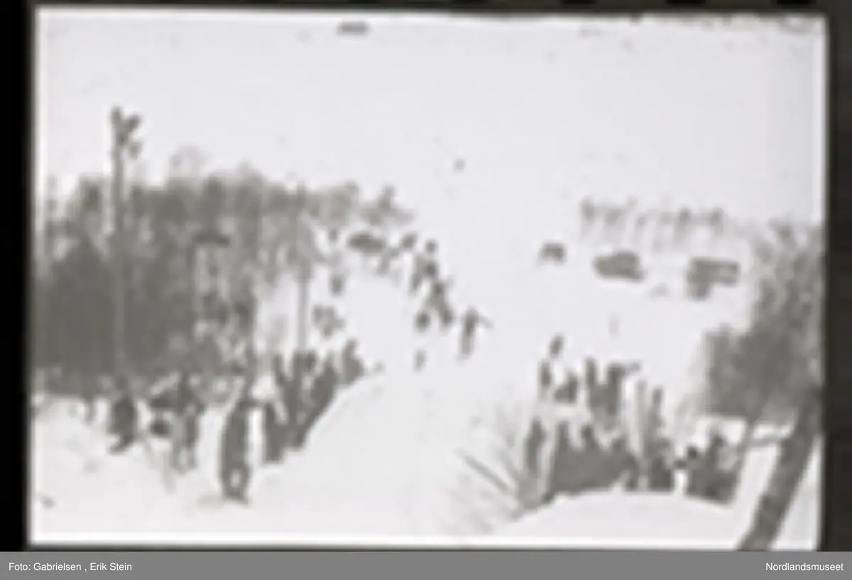 Fotografi av et skihopp som det hopper et barn med ski på seg fra mens noen kvinner og menn og barn med ski og skistaver og vinterklær og deltakernummer på seg som står og ser på barnet som hopper fra skihoppet vedsiden av skihoppet og lenger nede i bakken og tre barn som står i et utsiktstårn 
og ser på skihopperen og noen biler og busser som står parkert lenger nede i skibakken vedsiden av en vei som det vokser noen trær vedsiden av og man ser en skiløper ute i snømarken som har nettopp landet i snøen etter å ha hoppet fra skihoppet