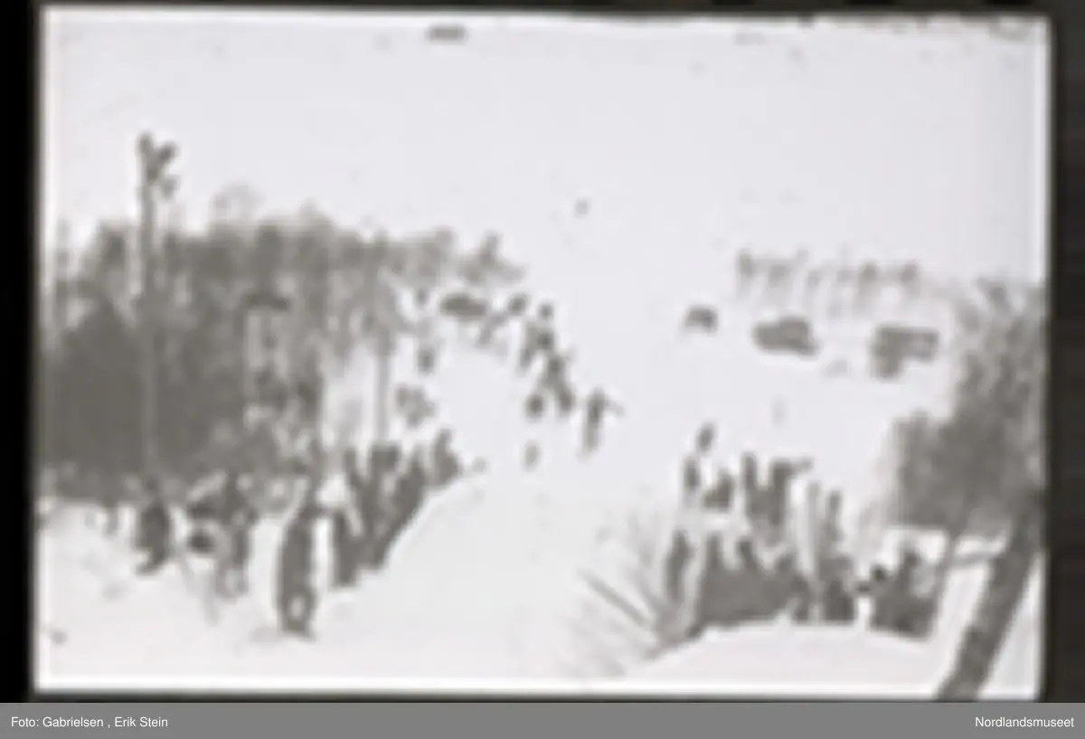 Fotografi av et skihopp som det hopper et barn med ski på seg fra mens noen kvinner og menn og barn med ski og skistaver og vinterklær og deltakernummer på seg som står og ser på barnet som hopper fra skihoppet vedsiden av skihoppet og lenger nede i bakken og tre barn som står i et utsiktstårn 
og ser på skihopperen og noen biler og busser som står parkert lenger nede i skibakken vedsiden av en vei som det vokser noen trær vedsiden av og man ser en skiløper ute i snømarken som har nettopp landet i snøen etter å ha hoppet fra skihoppet