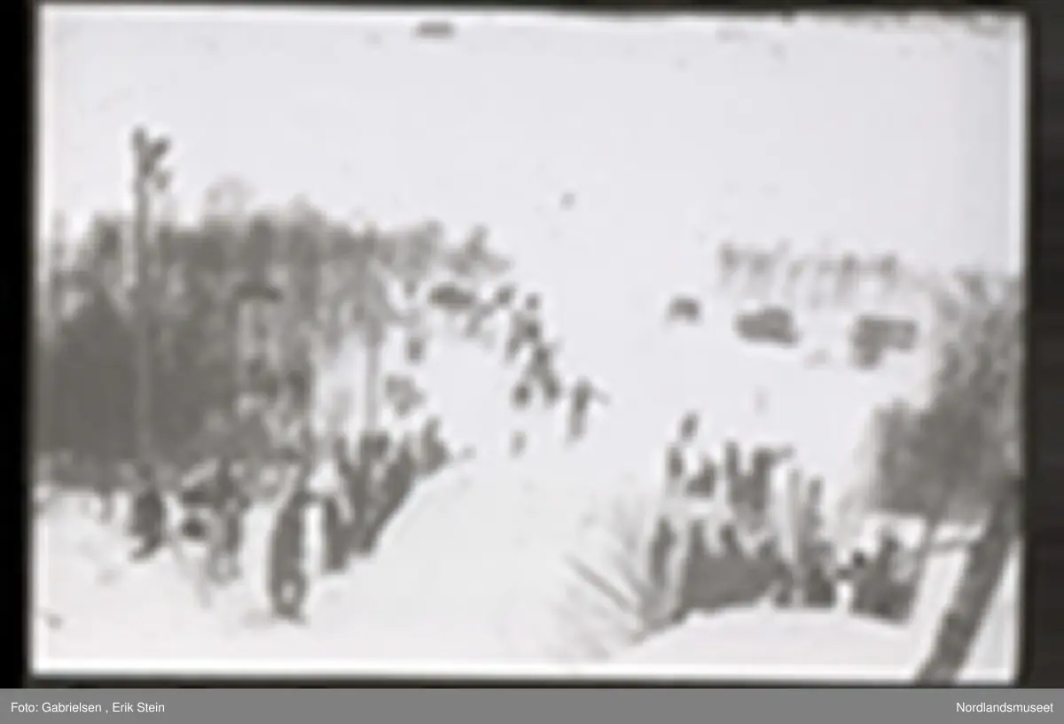 Fotografi av et skihopp som det hopper et barn med ski på seg fra mens noen kvinner og menn og barn med ski og skistaver og vinterklær og deltakernummer på seg som står og ser på barnet som hopper fra skihoppet vedsiden av skihoppet og lenger nede i bakken og tre barn som står i et utsiktstårn 
og ser på skihopperen og noen biler og busser som står parkert lenger nede i skibakken vedsiden av en vei som det vokser noen trær vedsiden av og man ser en skiløper ute i snømarken som har nettopp landet i snøen etter å ha hoppet fra skihoppet