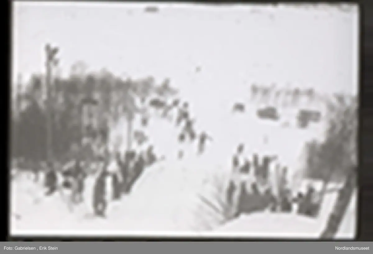 Fotografi av et skihopp som det hopper et barn med ski på seg fra mens noen kvinner og menn og barn med ski og skistaver og vinterklær og deltakernummer på seg som står og ser på barnet som hopper fra skihoppet vedsiden av skihoppet og lenger nede i bakken og tre barn som står i et utsiktstårn 
og ser på skihopperen og noen biler og busser som står parkert lenger nede i skibakken vedsiden av en vei som det vokser noen trær vedsiden av og man ser en skiløper ute i snømarken som har nettopp landet i snøen etter å ha hoppet fra skihoppet