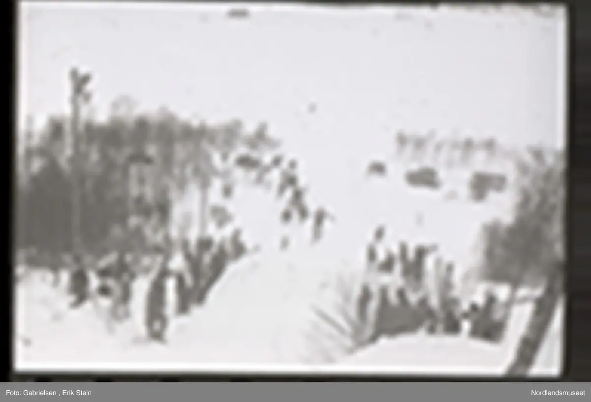 Fotografi av et skihopp som det hopper et barn med ski på seg fra mens noen kvinner og menn og barn med ski og skistaver og vinterklær og deltakernummer på seg som står og ser på barnet som hopper fra skihoppet vedsiden av skihoppet og lenger nede i bakken og tre barn som står i et utsiktstårn 
og ser på skihopperen og noen biler og busser som står parkert lenger nede i skibakken vedsiden av en vei som det vokser noen trær vedsiden av og man ser en skiløper ute i snømarken som har nettopp landet i snøen etter å ha hoppet fra skihoppet