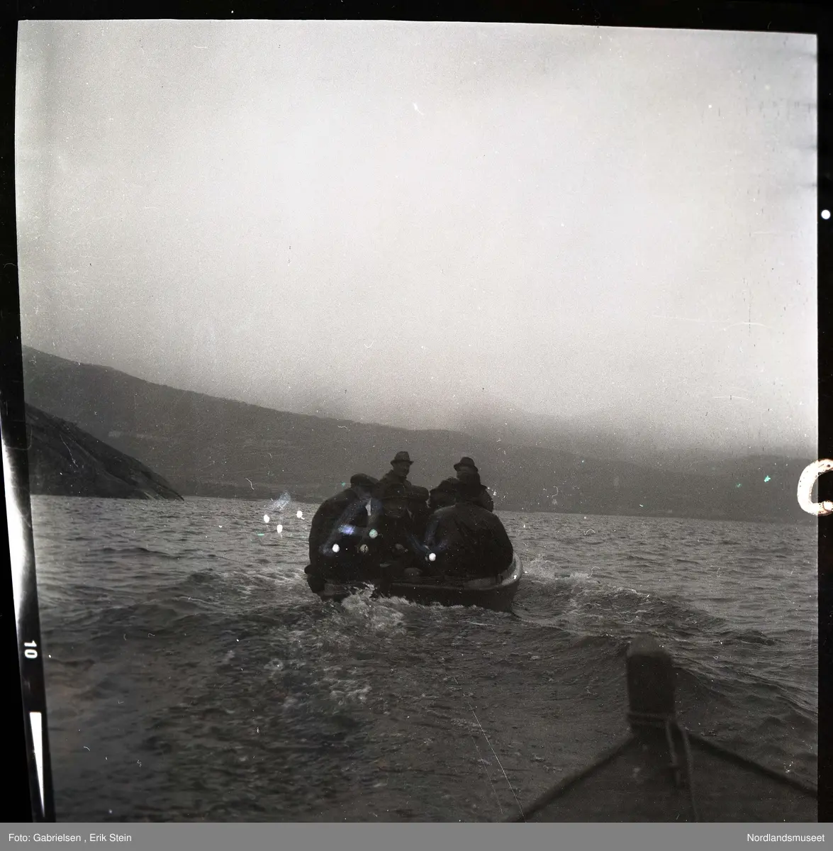 Fotografi av en motorbåt med fem menn som kjører ute på fjorden tatt fra en annen båt på fjorden bak motorbåten
med alle mennene eller en brygge til en kai som motorbåten kjører fra man ser at været er litt tåkete 
og man ser at alle mennene som sitter på motorbåten
er kledd i svarte jakker og hatter
og man ser noen berg som går ned til fjorden og
en li og et fjell på land på andre siden av fjorden