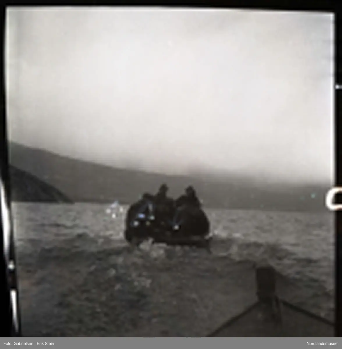 Fotografi av en motorbåt med fem menn som kjører ute på fjorden tatt fra en annen båt på fjorden bak motorbåten
med alle mennene eller en brygge til en kai som motorbåten kjører fra man ser at været er litt tåkete 
og man ser at alle mennene som sitter på motorbåten
er kledd i svarte jakker og hatter
og man ser noen berg som går ned til fjorden og
en li og et fjell på land på andre siden av fjorden