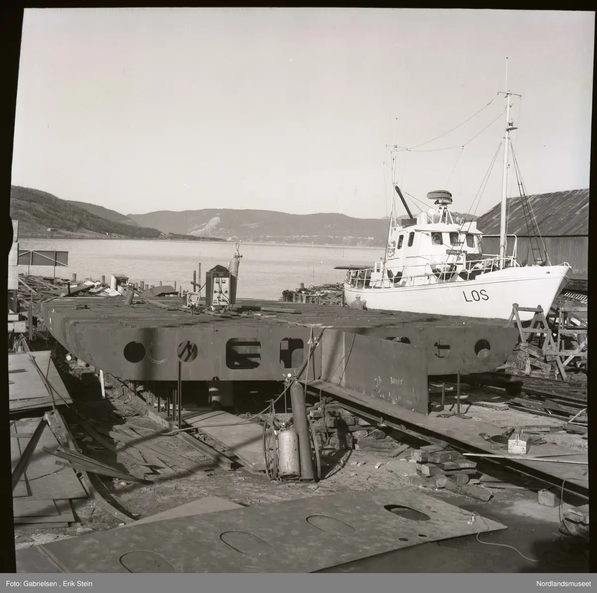 Fotografi av en båt under bygging på et båtbyggeri og verksted
vedsiden av en hvit båt som heter Los som står på noen stilasser som er inne for noen skader og skal repareres
og et båthus som står i nærheten av båten
som er inne for reparasjon og man ser en fjord
og noen lier med skoger og berg på andre siden av fjorden