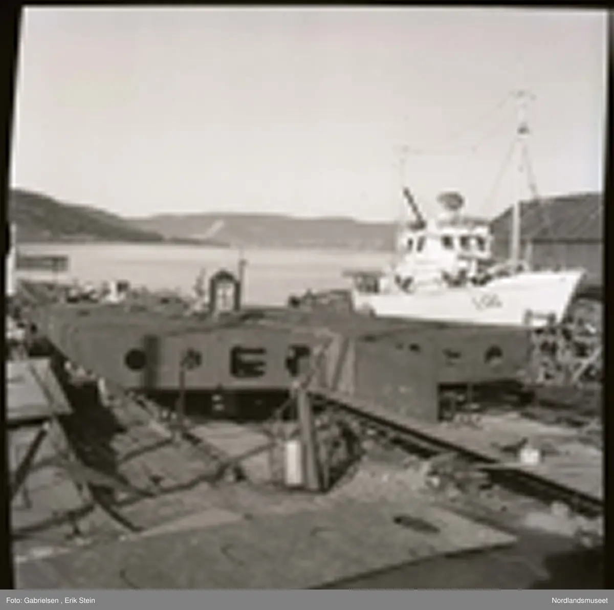 Fotografi av en båt under bygging på et båtbyggeri og verksted
vedsiden av en hvit båt som heter Los som står på noen stilasser som er inne for noen skader og skal repareres
og et båthus som står i nærheten av båten
som er inne for reparasjon og man ser en fjord
og noen lier med skoger og berg på andre siden av fjorden
