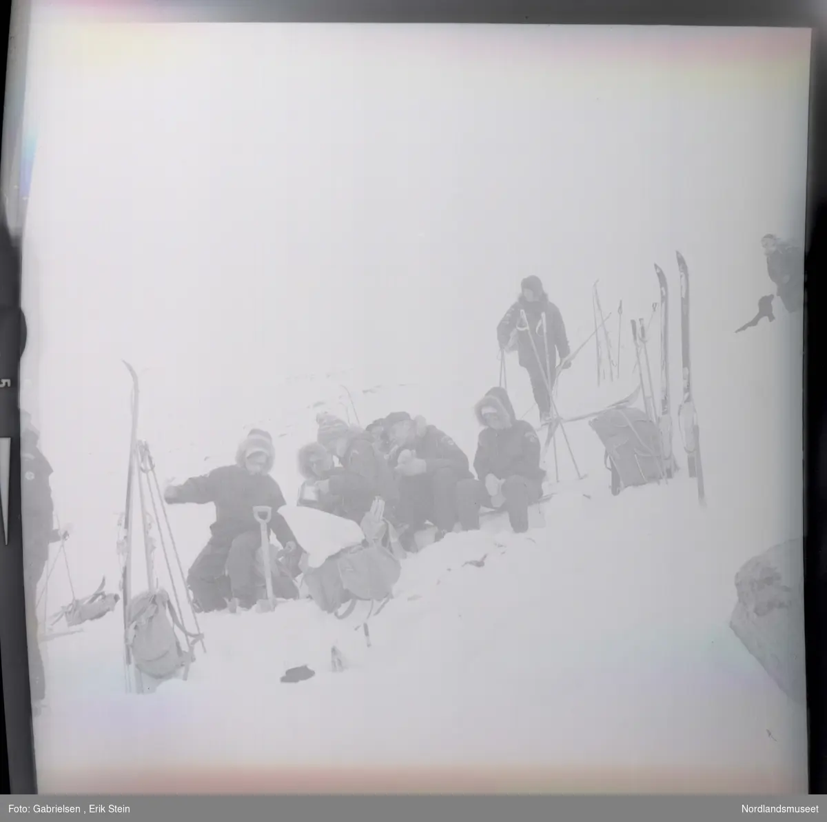 Fotografi av mange kvinner og menn kledd i vinterklær
som sitter på en snøbakke  på fjellet og graver etter mat og drikke i en tursekk som som ligger på snøen og en mann som går på ski på snøbakken over de og en kvinne som står på toppen av snøbakken vedsiden av en svart hund ¨
og man ser mange ski og skistaver og tursekker som står vedsiden av gruppen som er på skitur i fjellet 
og man ser litt av en mann som står på snøen vedsiden
av noen ski og skistaver og en tursekk som står i snøen