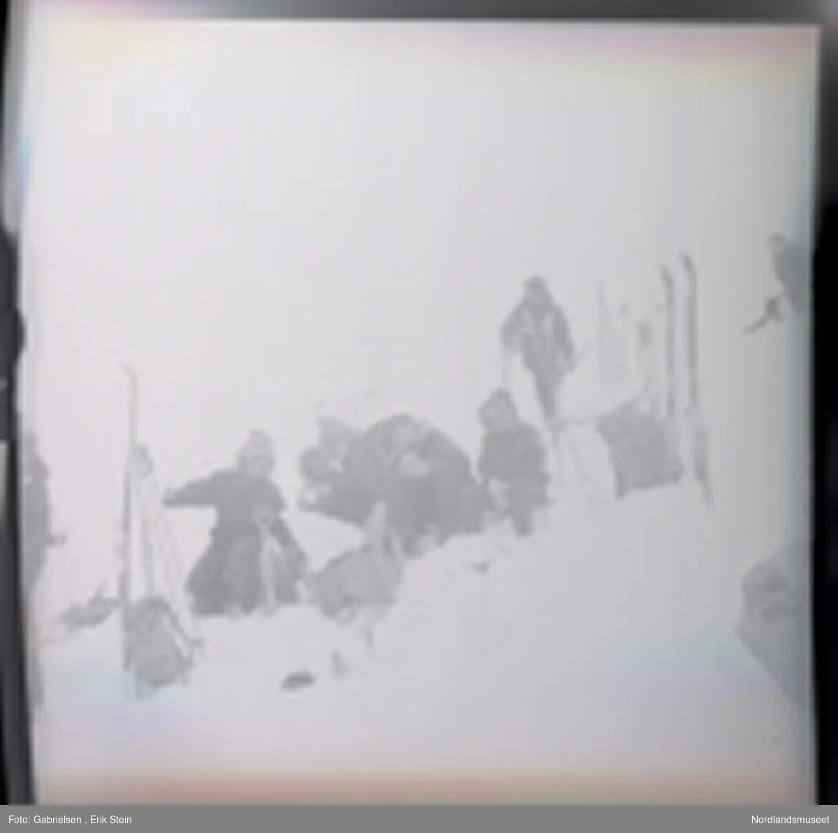 Fotografi av mange kvinner og menn kledd i vinterklær
som sitter på en snøbakke  på fjellet og graver etter mat og drikke i en tursekk som som ligger på snøen og en mann som går på ski på snøbakken over de og en kvinne som står på toppen av snøbakken vedsiden av en svart hund ¨
og man ser mange ski og skistaver og tursekker som står vedsiden av gruppen som er på skitur i fjellet 
og man ser litt av en mann som står på snøen vedsiden
av noen ski og skistaver og en tursekk som står i snøen