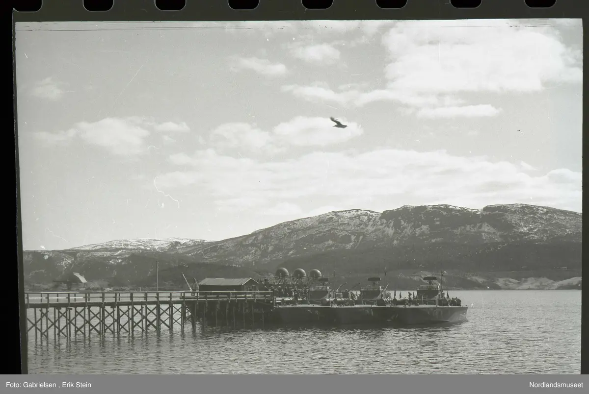 Fotografi av Fauskeviken som det går en lang brygge ut til 
som det står et båthus og en kran og noen krigsskip
som ligger fortøyd ved enden av bryggen som det går mange soldater ut fra og man ser mot Leivset og Finneid med
noen kaianlegg og anleggsområdet og 
man ser E6 motorveien som går over Leivset mot Rognan 
og noen fjell som det ligger litt snø på lenger borte