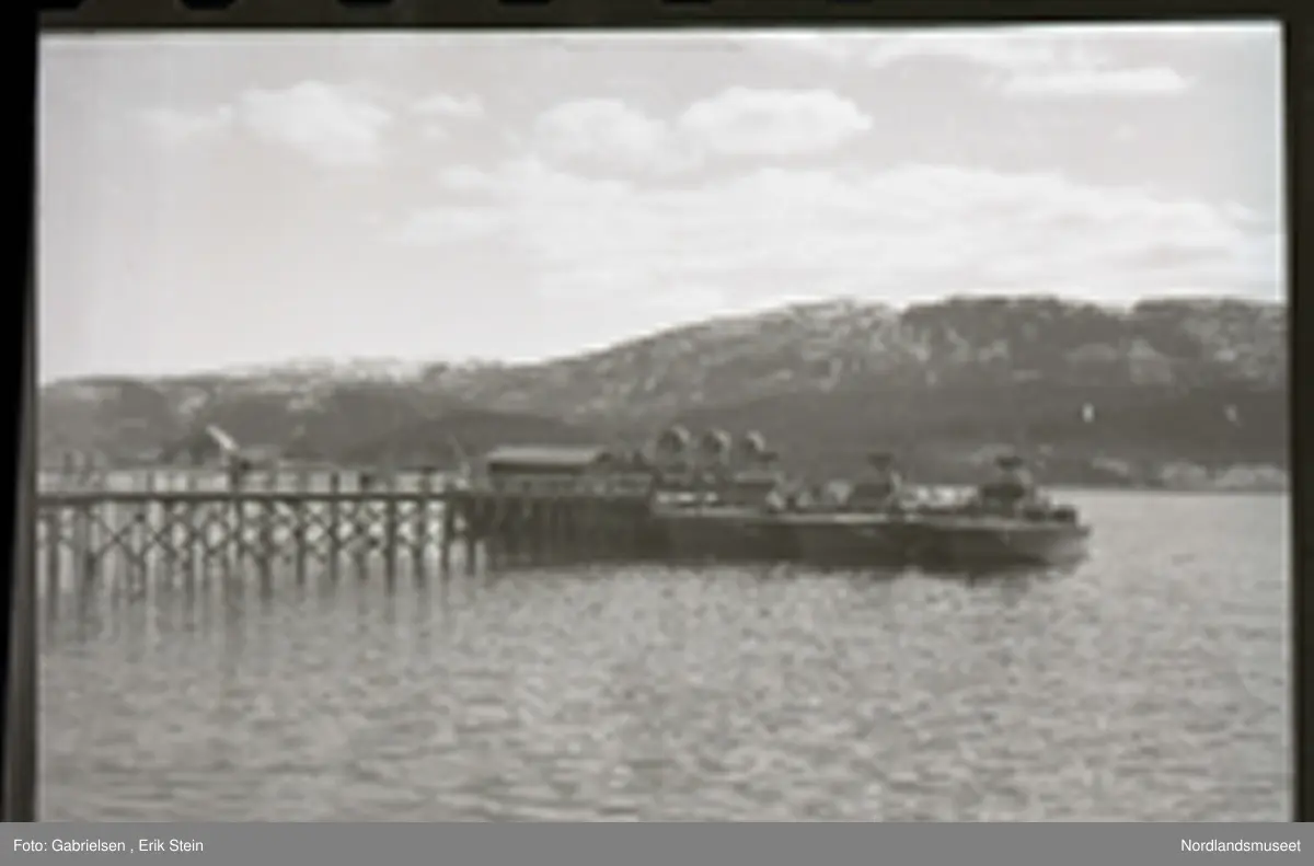 Fotografi av en lang brygge som går fra Fauskekaien 
ved Fauske sentrum ut til Fauskefjorden 
der man ser et båthus på og noen krigsskip
som ligger fortøyd ved bryggen og man ser en kran på bryggen vedsiden av båthuset på enden av bryggen 
og man ser mot Finneid og Leivset på andre siden av Fauskefjorden og man ser en strømstolpe som står 
på bryggen
