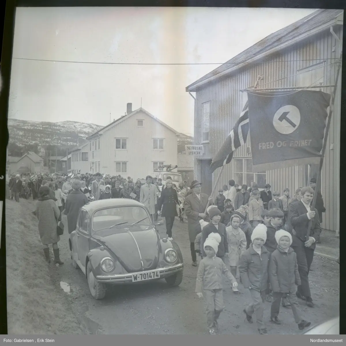 Fotografi av et 17 mai tog med mange kvinner og menn og barn som går i gatene i Fauske
forbi biler som står parkert på veien og noen bygninger 
i Fauske og Fauske lysverk sin bygning 
og en mann i toget som holder opp et flagg
og en annen mann som holder opp en plakat som det 
står fred og frihet med et hammersymbol