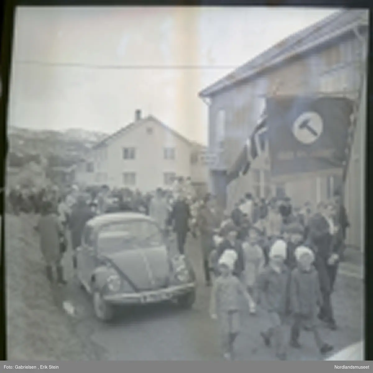 Fotografi av et 17 mai tog med mange kvinner og menn og barn som går i gatene i Fauske
forbi biler som står parkert på veien og noen bygninger 
i Fauske og Fauske lysverk sin bygning 
og en mann i toget som holder opp et flagg
og en annen mann som holder opp en plakat som det 
står fred og frihet med et hammersymbol