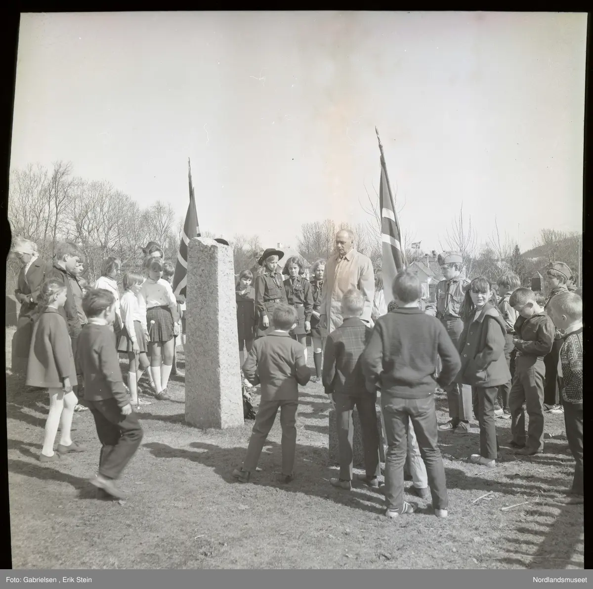 Fotografi av mange kvinner og menn og gutter 
og jenter som er finkledde 
som står og ser på en minnesmarkering 
av noen falne soldater på Fauske under andre verdenskrig
på 17 mai som en mann kledd i en hvit jakke
med slips som har lagt ned en krans på minnesmerket 
og noen soldater kledd i uniform ser på minnesmerket
og holder opp noen flagg på Fauske kirkegård