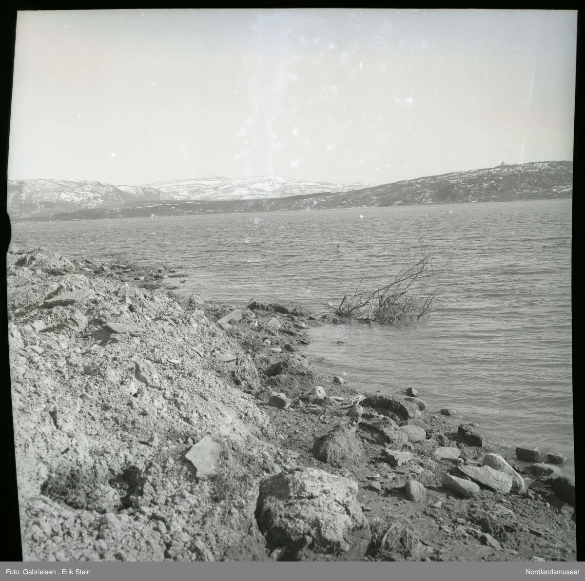 Fotografi av en fjære ved Lundfjorden 
med sand og mange steiner og noen trekvister som ligger
i fjorden ved fjæren og utsikt til Lundhalvøyen
og Erikstad som man ser mange lier med skoger
og kupplene til etteretningstjenestens 
anlegg på Fauske på toppen av Lundhalvøyen 
og man ser mange fjell som det ligger snø på 
mot Sulitjelma