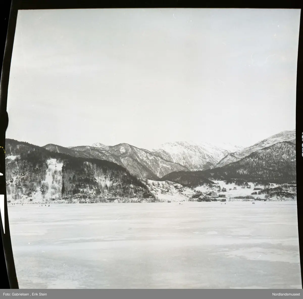 Fotografi av en fjord som er frosset til is som det går noen personer på og en vei på land som går mot noen hus og noen gårdsplasser og noen sjøbuer som står i fjæren ved fjorden og noen skoger som vokser i lier ovenfor husene og gårdsplassene og veien og fjell ovenfor liene som det ligger masse snø på