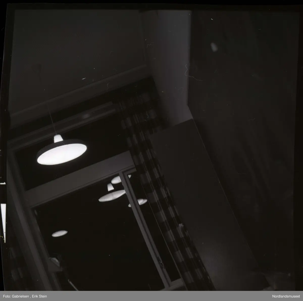 Fotografi av noen vinduer med gardiner i et klasserom 
i en skole og en tavle på veggen og noen lamper som henger
i taket som man ser i et speilbilde på vinduene i klasserommet