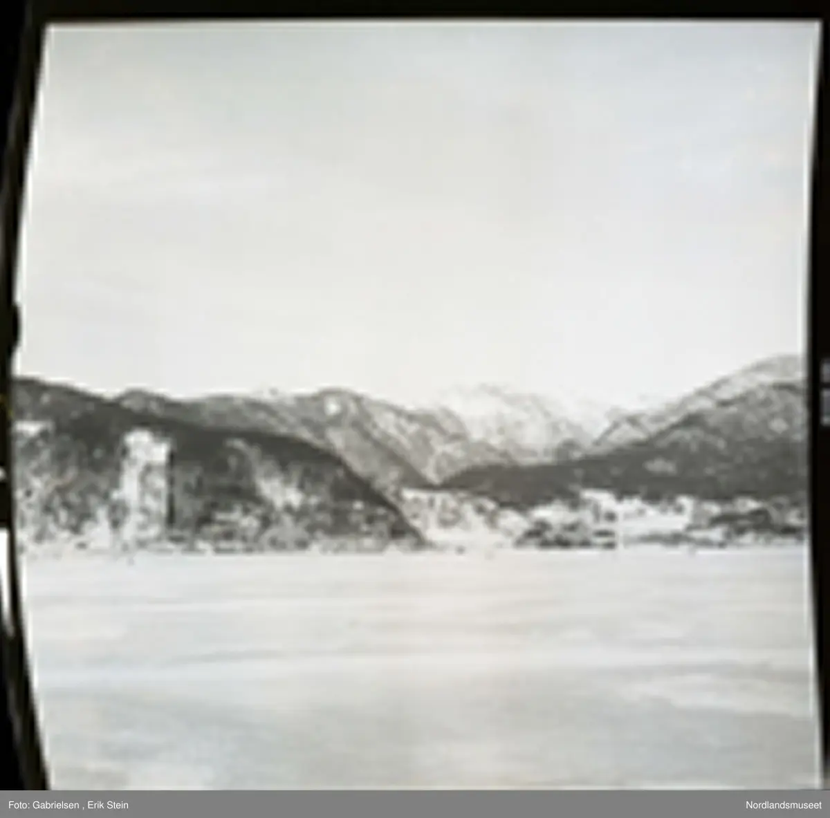 Fotografi av en fjord som er frosset til is som det går noen personer på og en vei på land som går mot noen hus og noen gårdsplasser og noen sjøbuer som står i fjæren ved fjorden og noen skoger som vokser i lier ovenfor husene og gårdsplassene og veien og fjell ovenfor liene som det ligger masse snø på