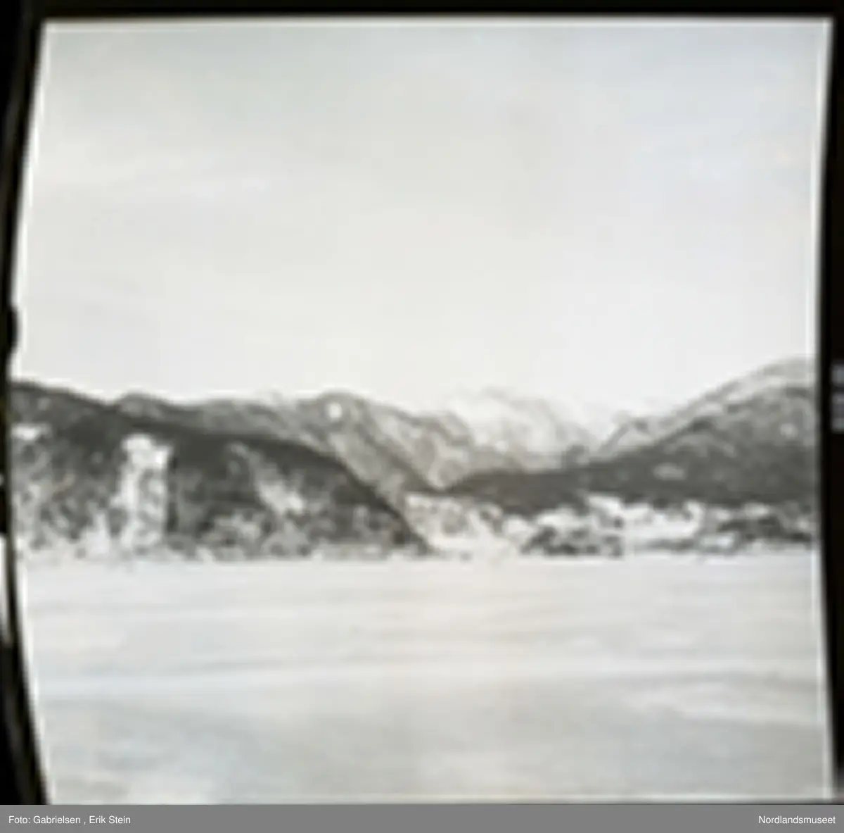 Fotografi av en fjord som er frosset til is som det går noen personer på og en vei på land som går mot noen hus og noen gårdsplasser og noen sjøbuer som står i fjæren ved fjorden og noen skoger som vokser i lier ovenfor husene og gårdsplassene og veien og fjell ovenfor liene som det ligger masse snø på