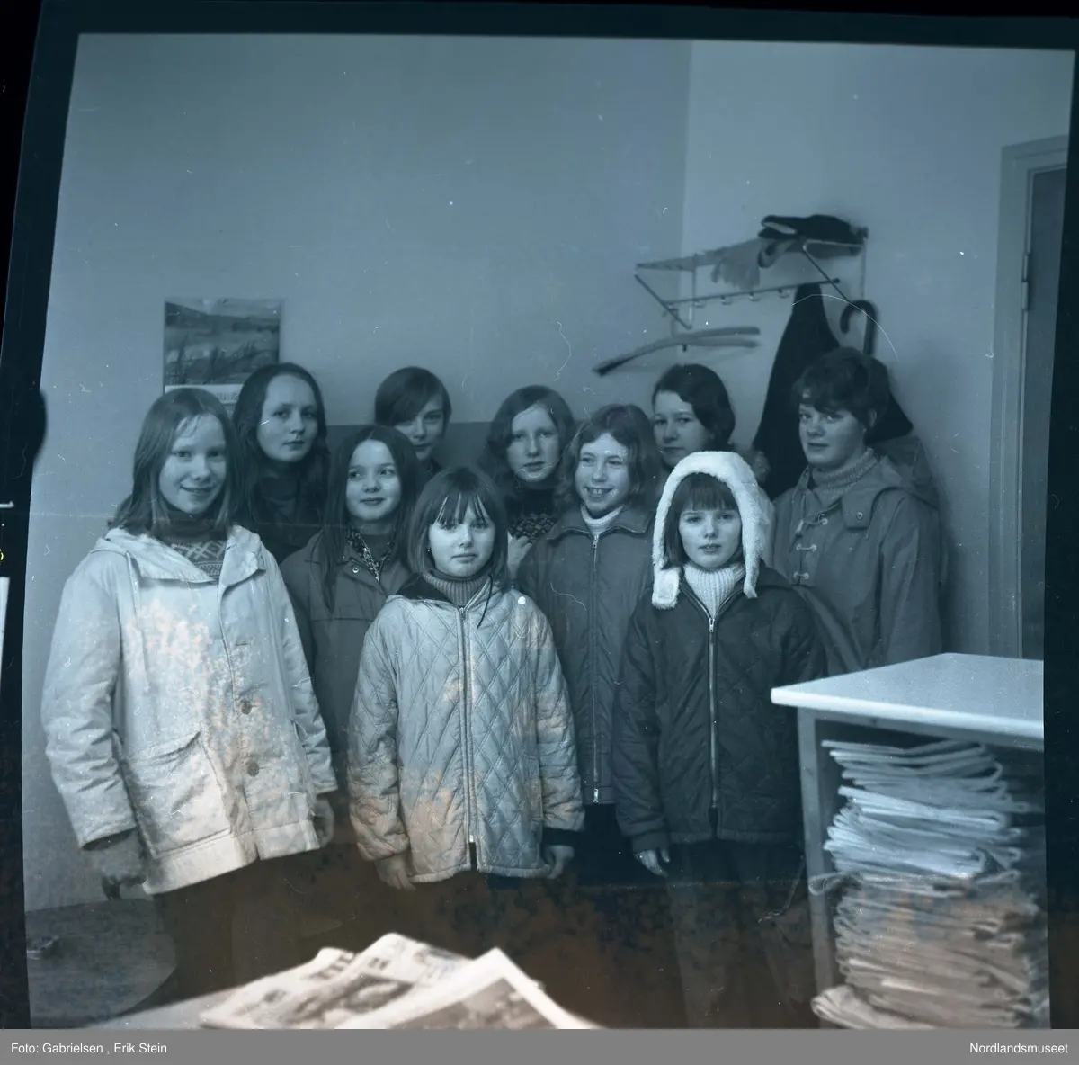 Fotografi av ti jenter kledd i vinterklær som står i et rom med 
et bord og en hylle som det ligger mange aviser på 
og en hylle som henger i veggen med et klestativ til 
og henge klærene sine på som det henger en jakke på 
og ligger et skjerf og et par votter og en kalender
som henger på veggen og en dør som går ut av rommet