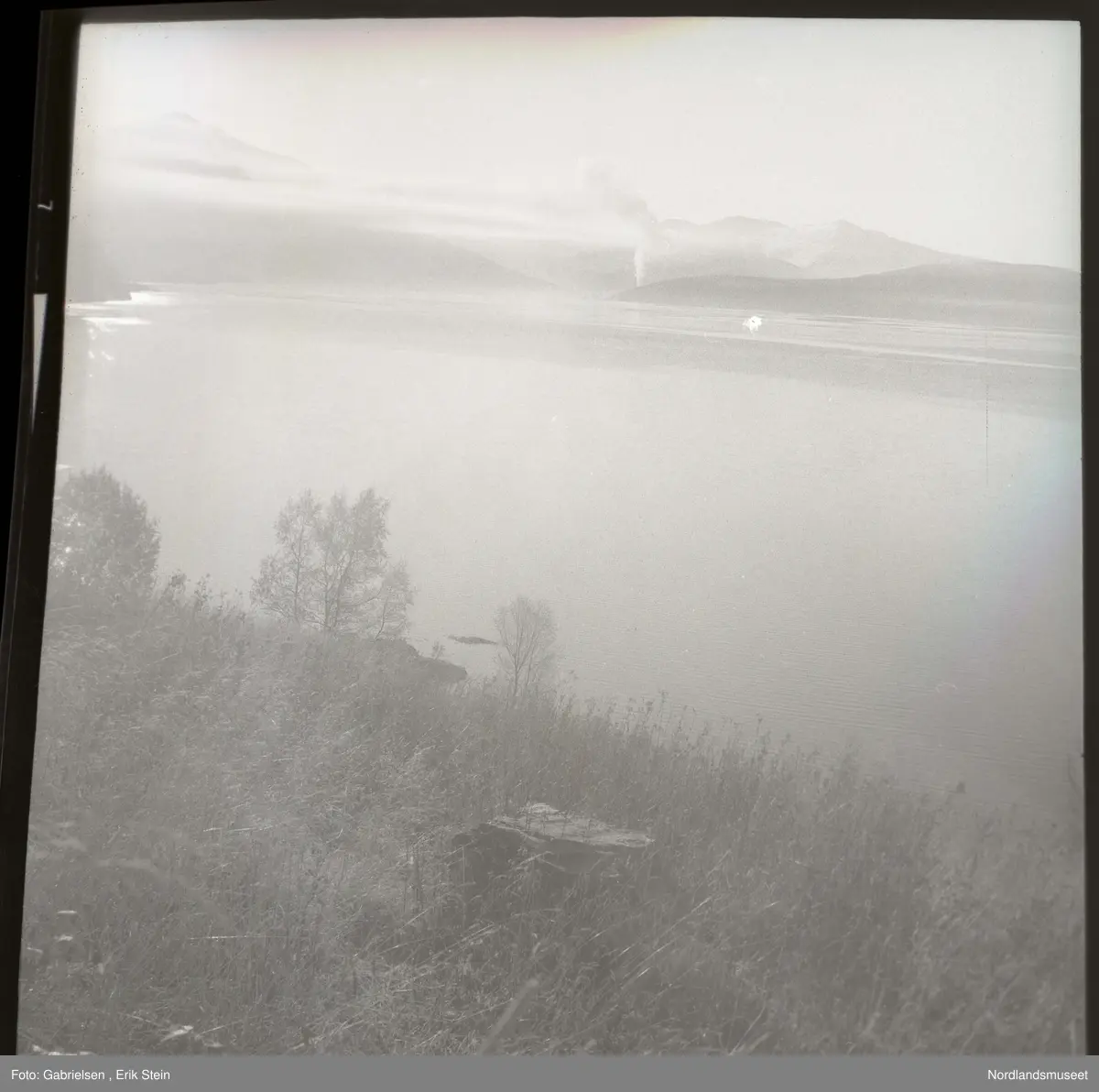 Fotografi av en bakke som det bokser mange trær og busker
og gress på som går ned til en stor fjord som man på 
andre siden på land ser mange skogsområder på og noen lier 
og noen fjelltopper lenger unna og man ser en stor stein
som ligger i bakken som går ned til fjorden og noen skyer
på fjellene lenger unna