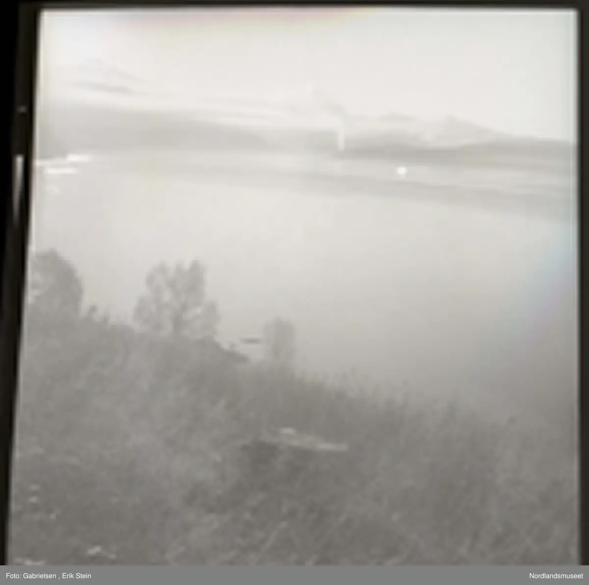 Fotografi av en bakke som det bokser mange trær og busker
og gress på som går ned til en stor fjord som man på 
andre siden på land ser mange skogsområder på og noen lier 
og noen fjelltopper lenger unna og man ser en stor stein
som ligger i bakken som går ned til fjorden og noen skyer
på fjellene lenger unna