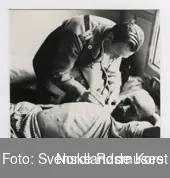 Fotografi av en doktor kledd i en uniform 
som måler pulsen til  en krigsfange som ligger 
på en seng inne i sykehusbrakken i Klungset leir
og holder skjorten til krigsfangen oppe som man kan 
den bare magen til krigsfangen som er liten og tynn
som man kan se skjellettet til krigsfanget 
og man ser at et vindu vedsiden av sengen som krigsfangen
ligger i