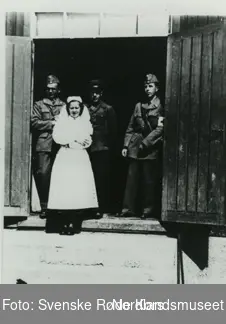 Fotografi av en sykepleier som står med to leger
og en soldat kledd i uniformer på inngangen til Sykehusbrakken i Klungset leir
med en trapp og en dobbeldør som står åpen
og fire tynne vinduer over dobbeldøren