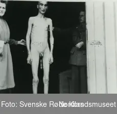 Fotografi av en naken tynn utsultet krigsfange som man kan se skjelettet på 
som står vedsiden av en lege og en sykepleier 
i en inngang til et sykehus