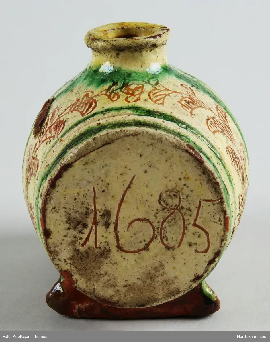 Flaska i form av liggande tunna med dekor av gröna bårder som markerar tunnbanden och blad ristade i den gulvita engoben. På ena kortsidan märkt "1685". 
Se Bonge-Begengren 1994, s 25.