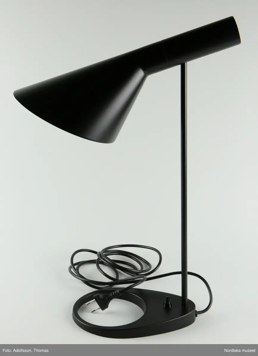 a) Bordslampa av svart pulverlackerat stål. Smal cylindrisk skärm vidgar sig nedåt till asymmetrisk trumpetform. Smal cylindrisk fot fäst i närmast oval fot med runt utsparat hål. Strömbrytare på foten, svart filt under densamma. Sladd av svart plast med stickpropp. Lampfattning E27 med b) isittande glödlampa. 

Mindre skrapmärken framtill på foten.
/Maria Maxén 2021-12-20