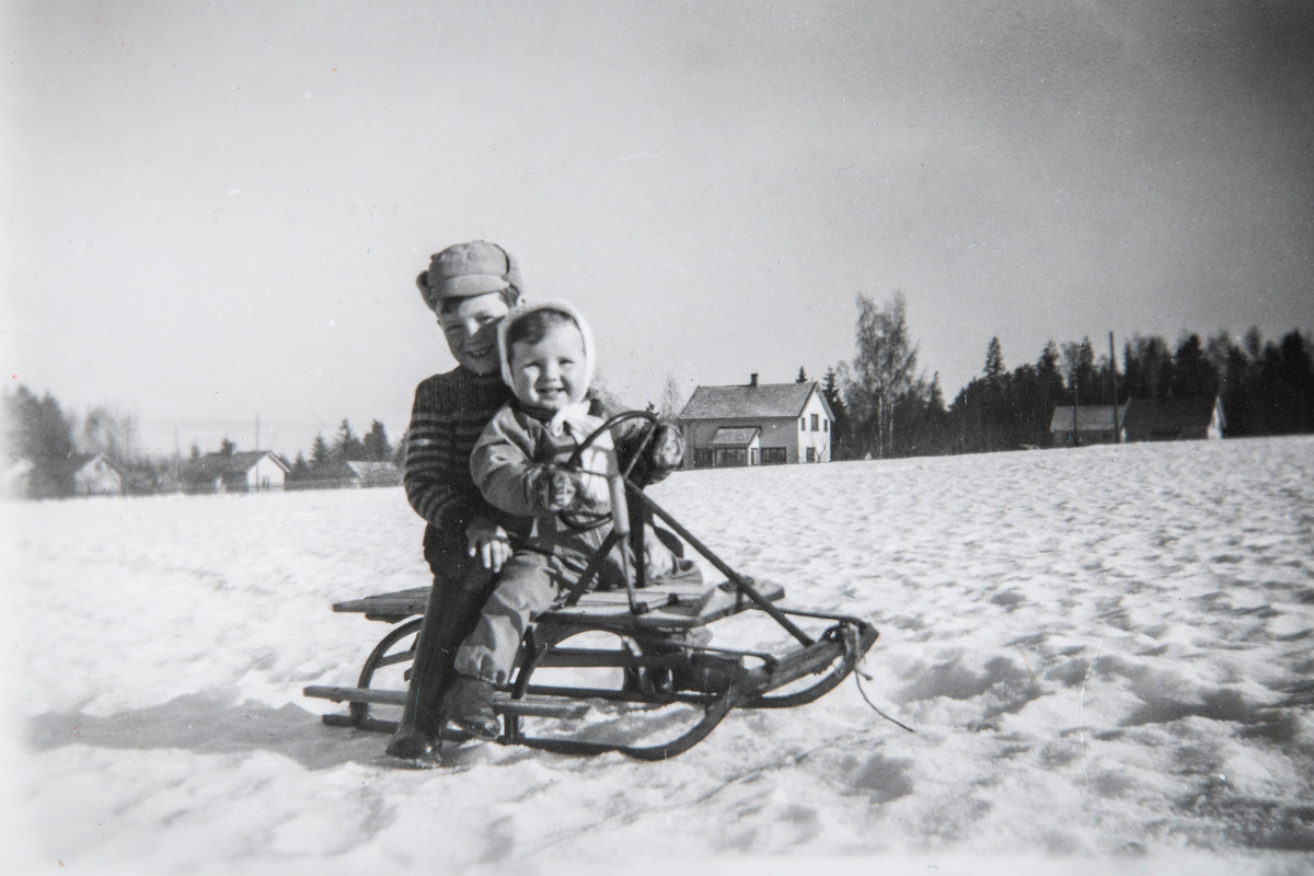2 barn sittende på en rattkjelke. Edgar Furulund (f. 1952) bakerst og Trond Furulund (f. 1957) foran. Huset i bakgrunnen ble eid av "Windhoel-familien". Bildet er fra ca 1959. Vinter, snø.