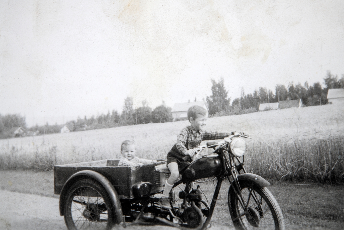 Edgar Furulund (f. 1952) prøver farens tre-hjuling. Trond Furulund (f. 1957) sitter på lastplanet. Bildet er tatt i Basterudvn. 20,  i 1958. Trehjuls motorsykkel. Vare motorsykkel.