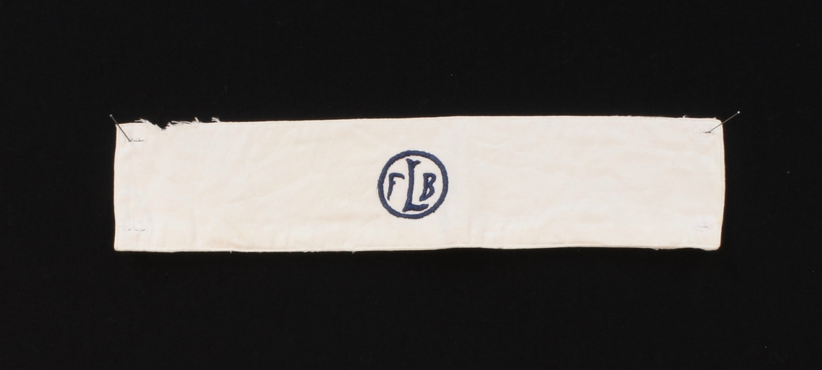 Vit armbindel med blå broderad text "FLB" inom cirkel. Två knapphål i var ände.
Sliten. På baksidan fastsydd märklapp med initialerna "B.H".