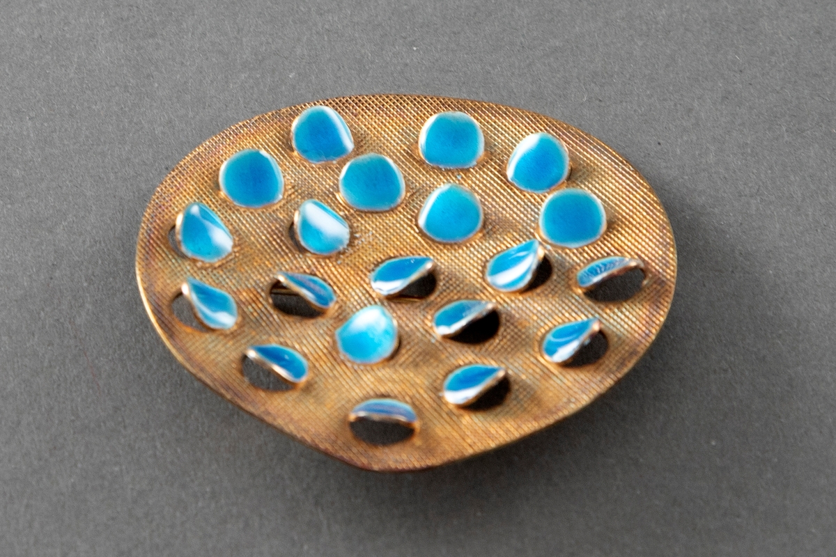 Brosje bestående av en tilnærmet rund plate i forgylt sølv med rutemønstret overflate og halvgjennombrutte, runde flak dekket med turkis emalje som åpner seg utover.