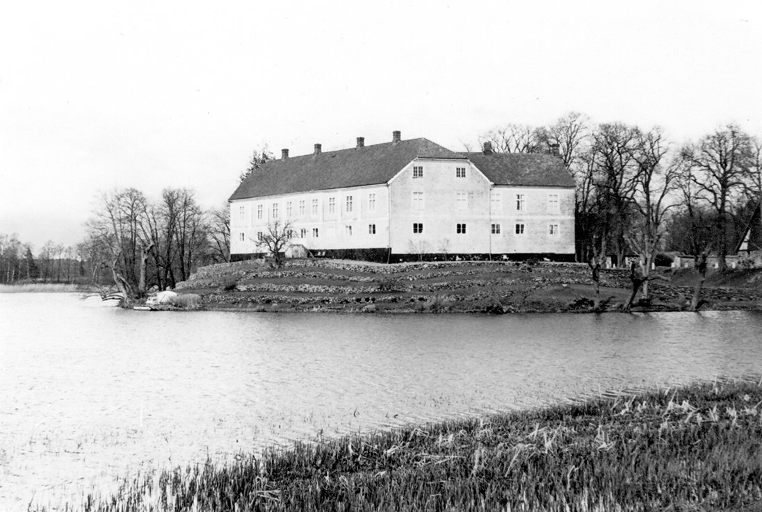 Vallens slott vid Storsjön, strax norr om orten Våxtorp, sett från sydväst. Byggnaden ligger på en liten holme med numera torrlagd vallgrav mot fastlandet (till höger i bild). Godsets historia kan följas tillbaka till 1300-talet och i manbyggnaden ingår rester av medeltidsborgen som tidigare stod här. Nuvarande utformning med en klassicerande fasad med pilastrar på övervåningen tillkom 1801, då slottet återuppbyggdes efter en brand året innan. Från att ha varit en fyrlängad borg, med torn över porten till vindbryggan, består det gulputsade slottet numera av en lång länga åt väster sammanbyggd med två kortare flyglar, i trubbiga vinklar, i norr och söder. Marken är flackt terrasserad.
(Se även bildnr E2686, F4511)