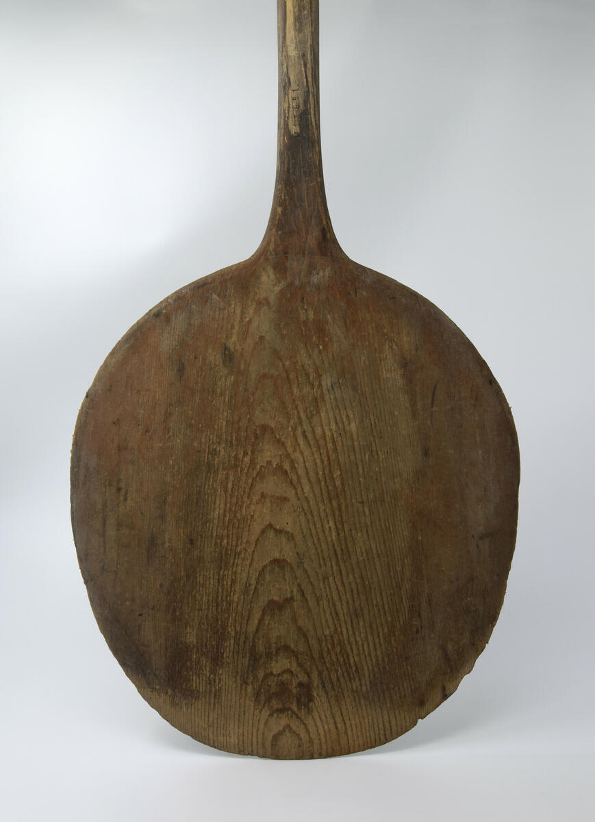 Brödspade, av björkträ gjord i ett enda stycke. Oval skiva med långt skaft. Skivans storlek 0,36 x 0,40 m. Nött länst hela skaftet och i toppen av skivan.