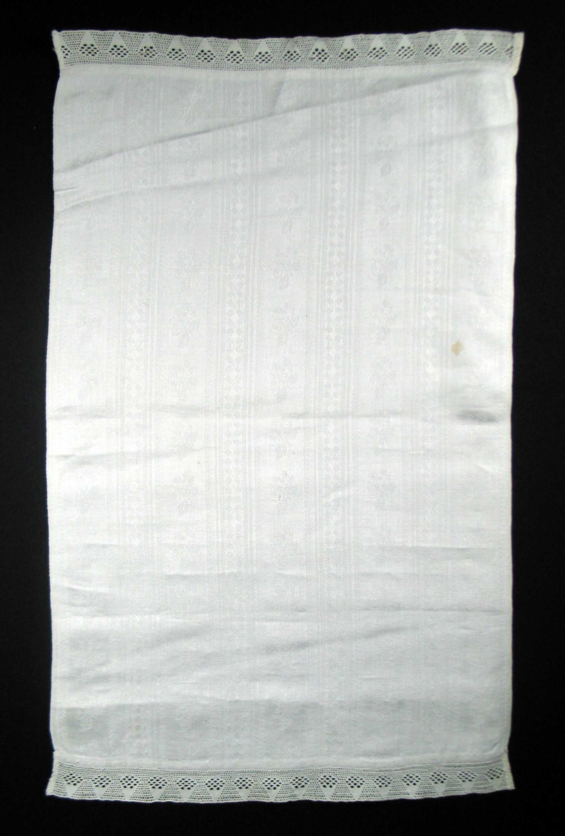 Hvit linduk med blonde påsydd i begge ender. Vevbredden er 45,5 cm. Lintøyet er stripete med border i lengderetningen. 4,5 rapport i bredden. Mellom, fast, rettvinklet oppbygd mønster med kryssfigurer, 5,5 bred er et felt med blad-og blomstmotiv hvor alle vender samme vei.