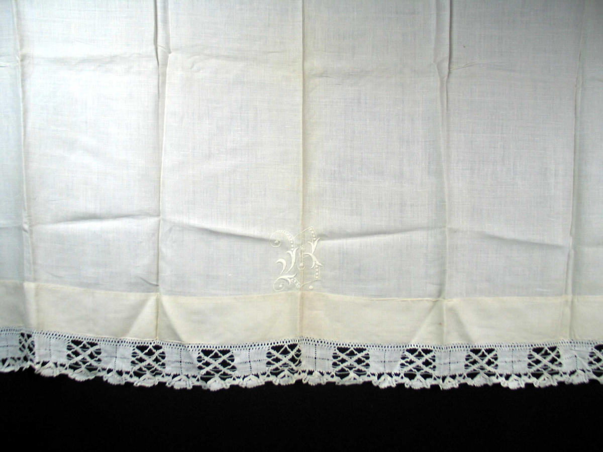 Hvit linduk med påsydd blonde nederst. Duken er sydd sammen av to stykker. Jare i ene siden, faldet i den andre. Midt på duken like over bred fald er det et tykksømsmonogram samt kontursting i hvitt.
