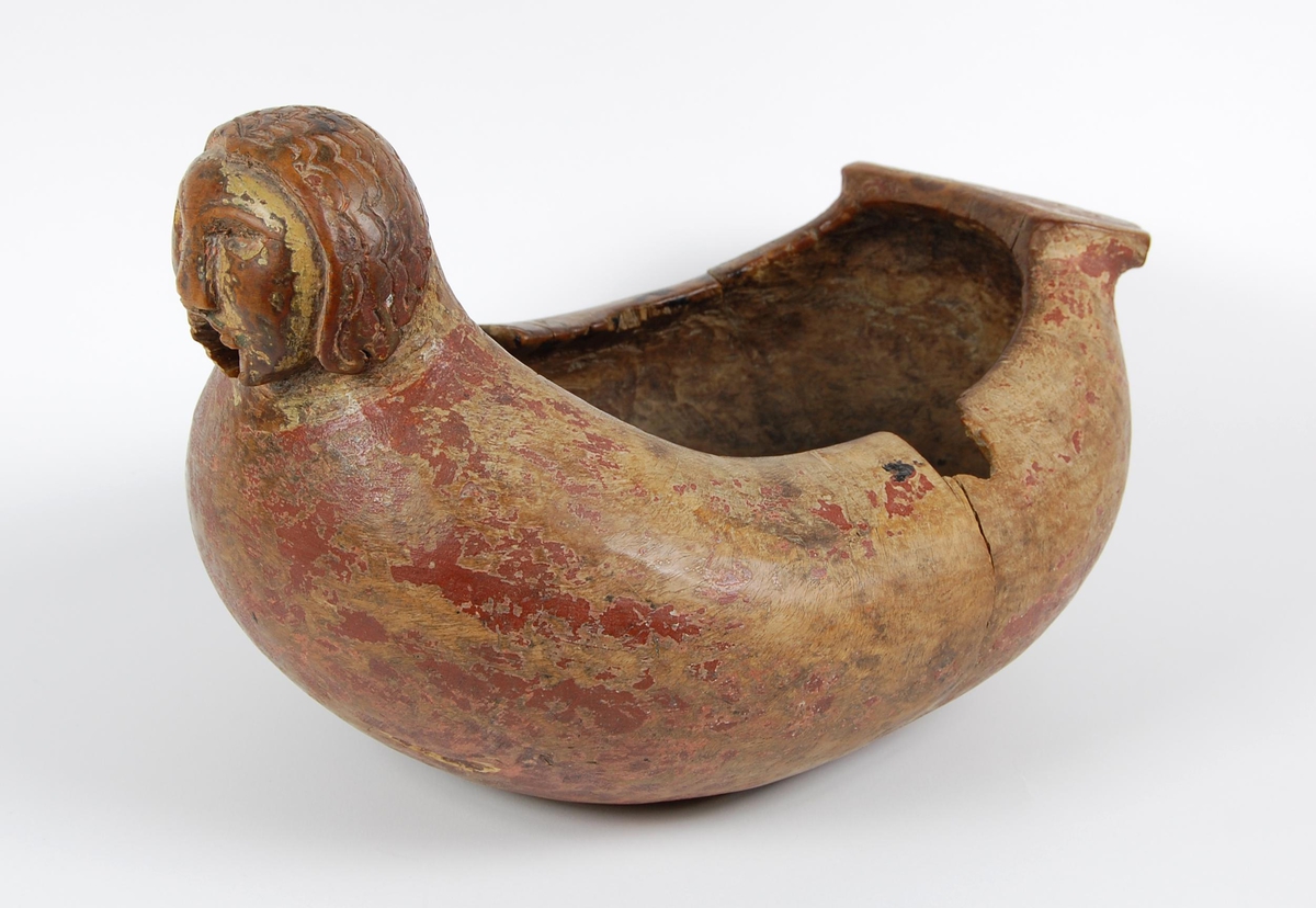 Trøys av bjørk. Oval form. Utskåret mannshode med munnåpning. Håndtak med hakkformete utskjæringer og innskrift. Svarte bekklumper i bunnen.